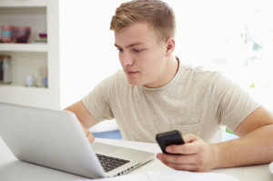 Teenage Boy Sending Text Message Whilst Studying On Laptop